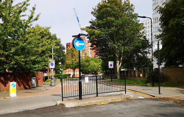 The barrier is a metal railing in the foreground; beyond it stand trees and high-rise tower blocks